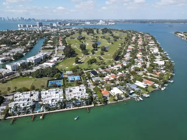 High-angle aerial photograph of a luxury real estate island community, showcasing waterfront houses, boat docks, multi-story condos, tennis courts, and a central green golf course surrounded by ocean water under a clear blue sky.