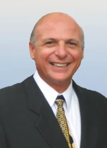 Professional headshot portrait of real estate agent Bob D'Andrea smiling, wearing a black suit jacket, white collared dress shirt, and a patterned gold tie.