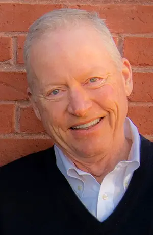 Professional headshot portrait of real estate agent Cash Wiseman smiling, wearing a dark sweater over a white collared shirt against a red brick wall background.