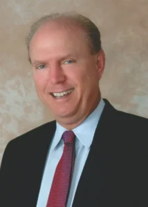 A professional headshot portrait of real estate agent John McWilliams smiling, wearing a black suit, light blue shirt, and red patterned tie against a neutral studio background.