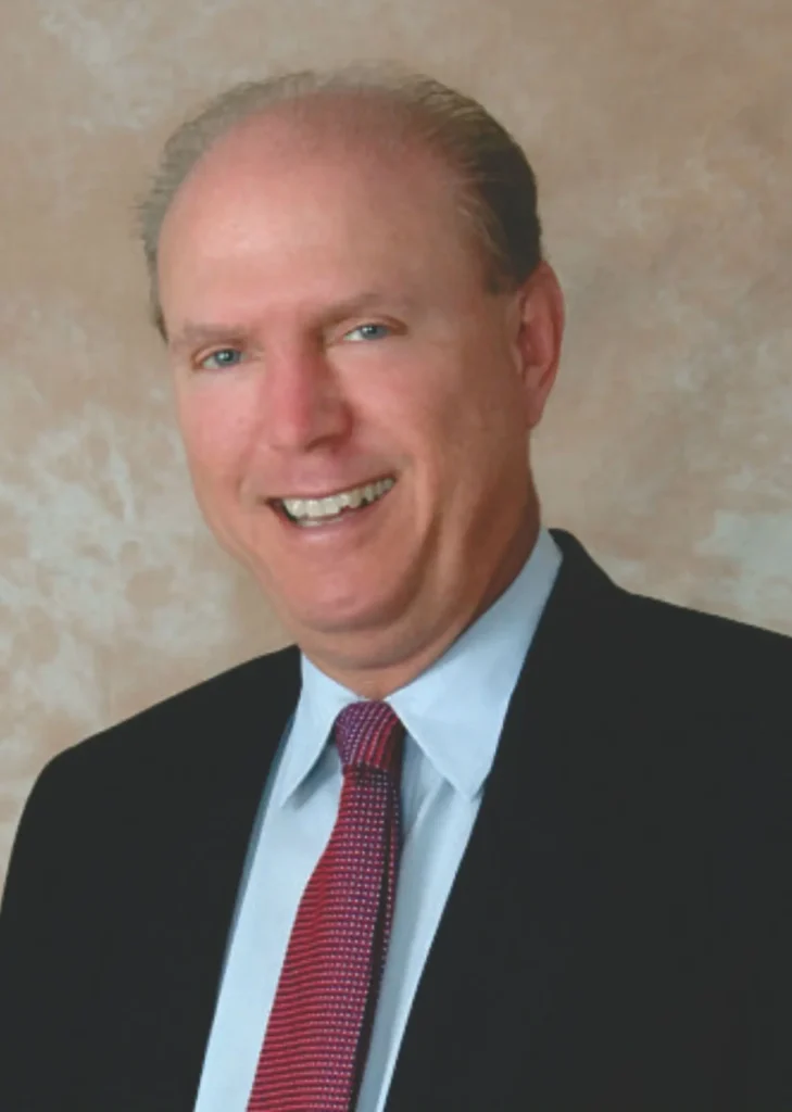 A professional headshot portrait of real estate agent John McWilliams smiling, wearing a black suit, light blue shirt, and red patterned tie against a neutral studio background.