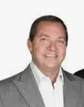 Professional headshot portrait of real estate agent Mark Miller smiling, wearing a grey suit jacket and white collared dress shirt against a white background.