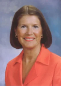 Professional headshot portrait of real estate agent Nancy Gurry smiling, wearing a coral orange blazer jacket against a mottled blue background.