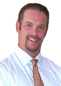 Professional headshot portrait of real estate broker Tom Buckley smiling, wearing a striped dress shirt and patterned gold tie against a white background.