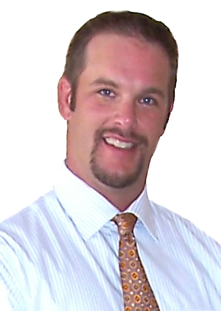 Professional headshot portrait of real estate broker Tom Buckley smiling, wearing a striped dress shirt and patterned gold tie against a white background.